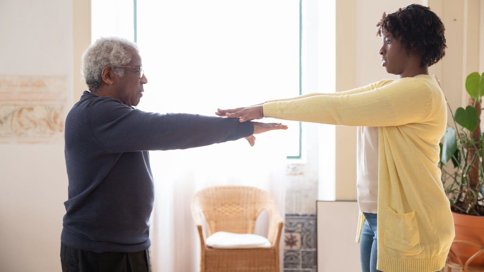 Homem idoso, em pé na sala de casa, estende os braços à frente acompanhando o movimento de uma mulher que o orienta.